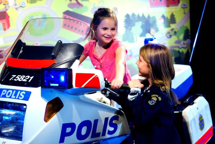 Två barn, 7-åriga flickor, leker i Polismuseets barnutställning. Den ena sitter på polismotorcykeln och den andra står bredvid med en polisjacka i barnstorlek på sig.